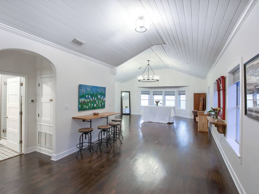 A spacious room with a high ceiling, wooden flooring, a dining area with stools, and a large painting on the wall.