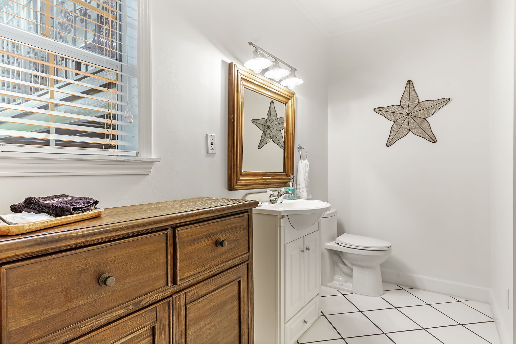 A bathroom with a wooden vanity, white sink, and toilet. There is a starfish decoration on the wall and a window with blinds.