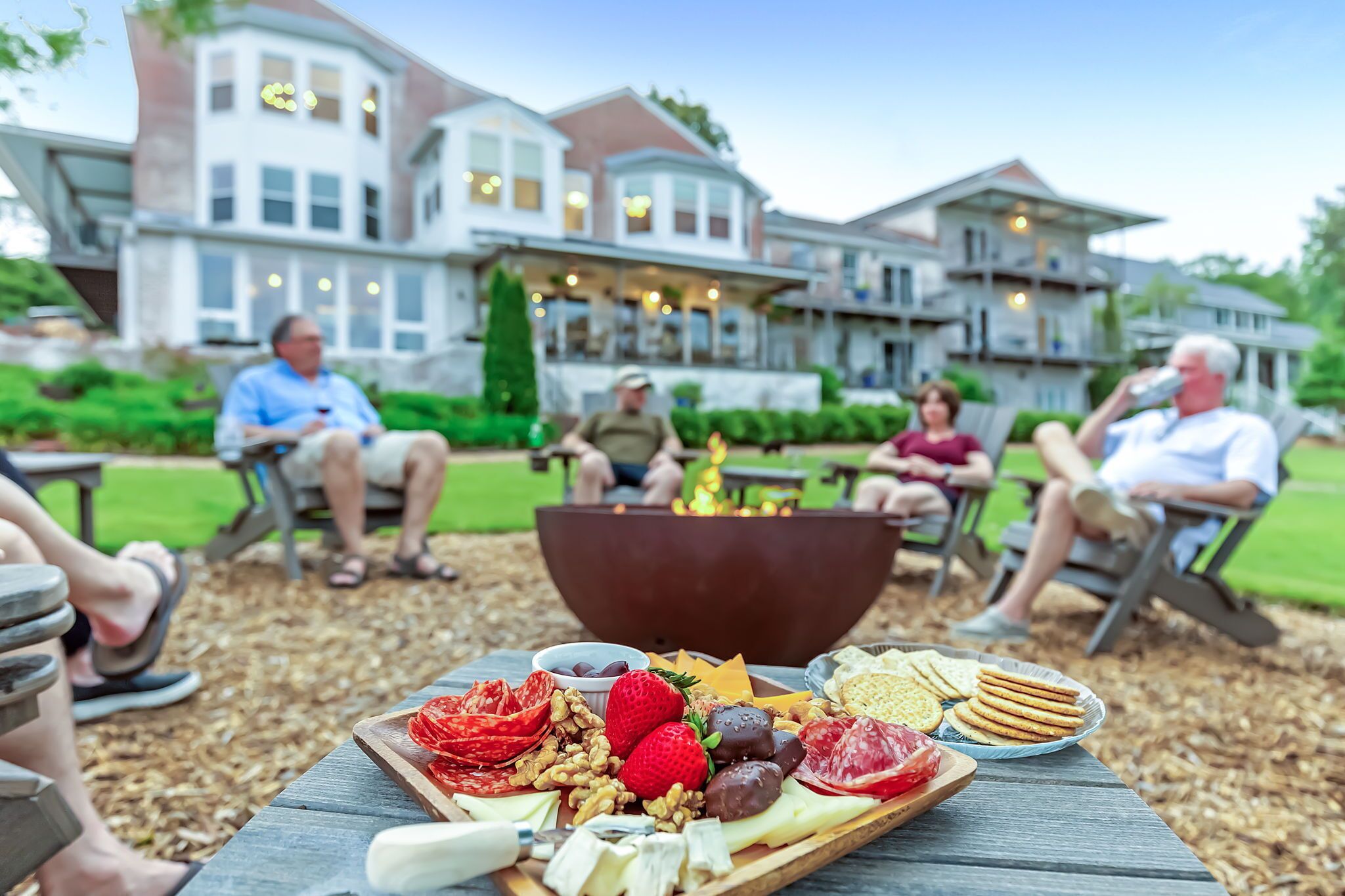 A group of people seated outdoors at a picnic table with a variety of cheeses, fruits, and crackers.