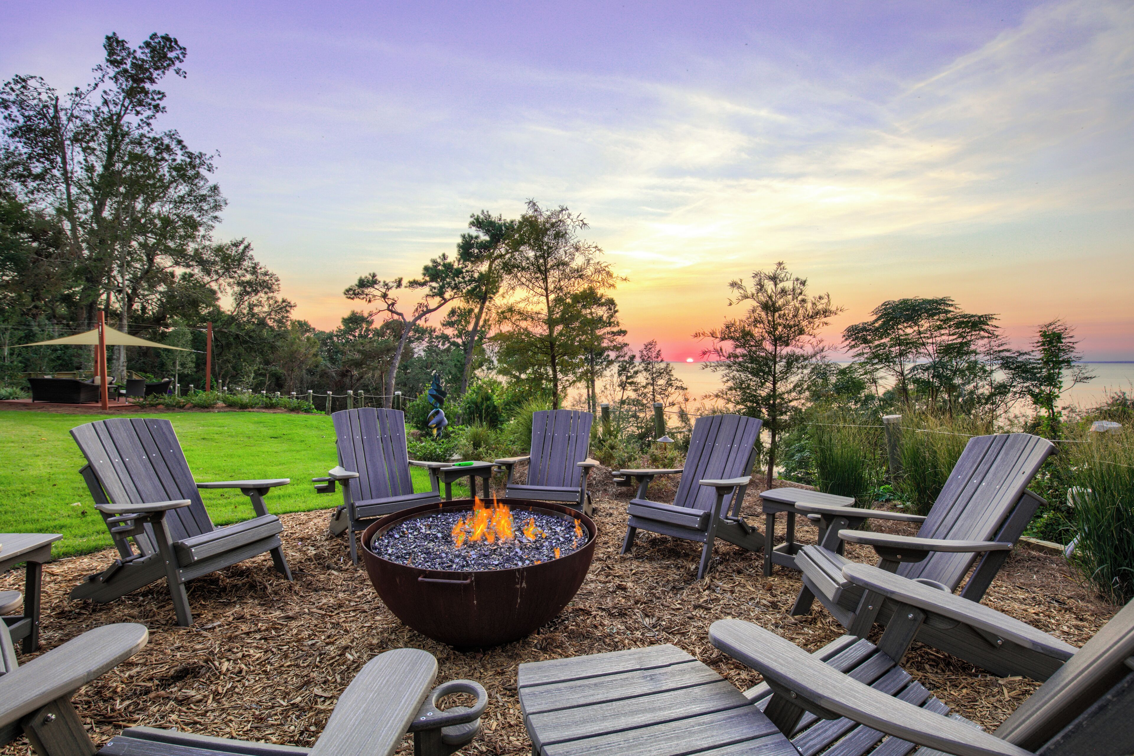 A fire pit with chairs and tables in a garden setting, overlooking a body of water and trees.