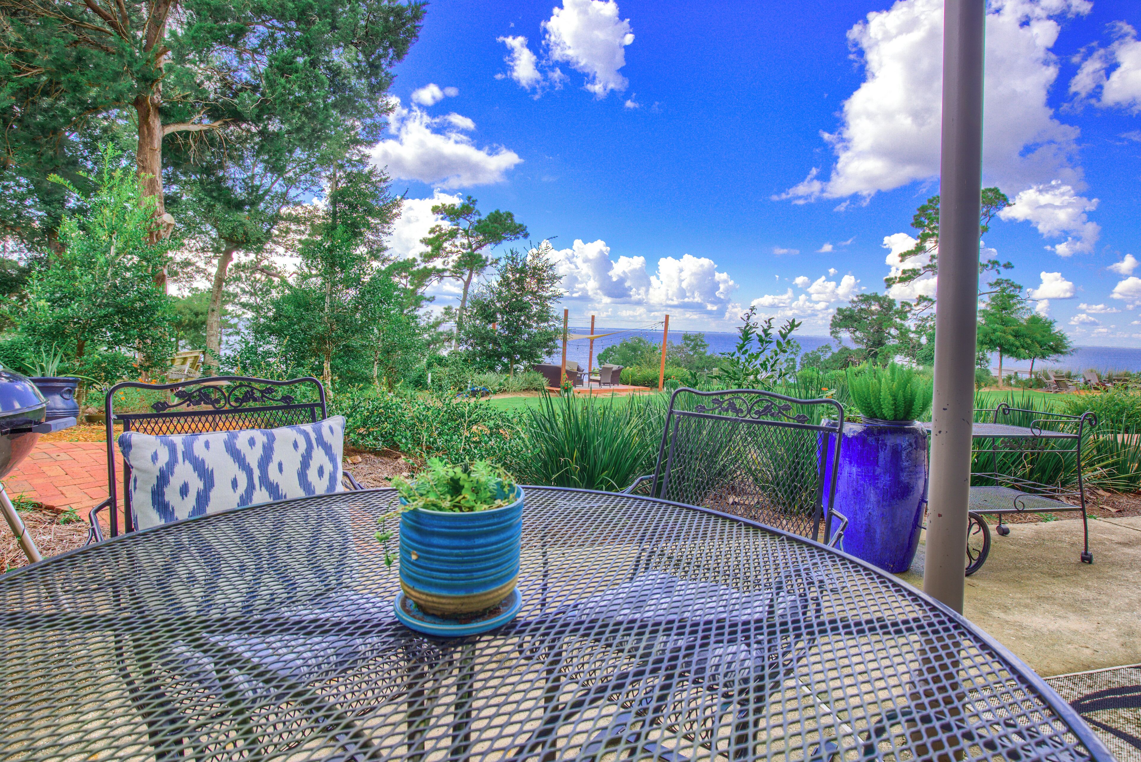 A metal table with a blue planter, surrounded by metal chairs with patterned cushions, set in an outdoor garden with trees and a clear sky.