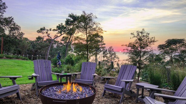 A fire pit with chairs and tables in an outdoor setting with a view of the sunset over water. - Jubilee Suites Boutique B&B (Fairhope)