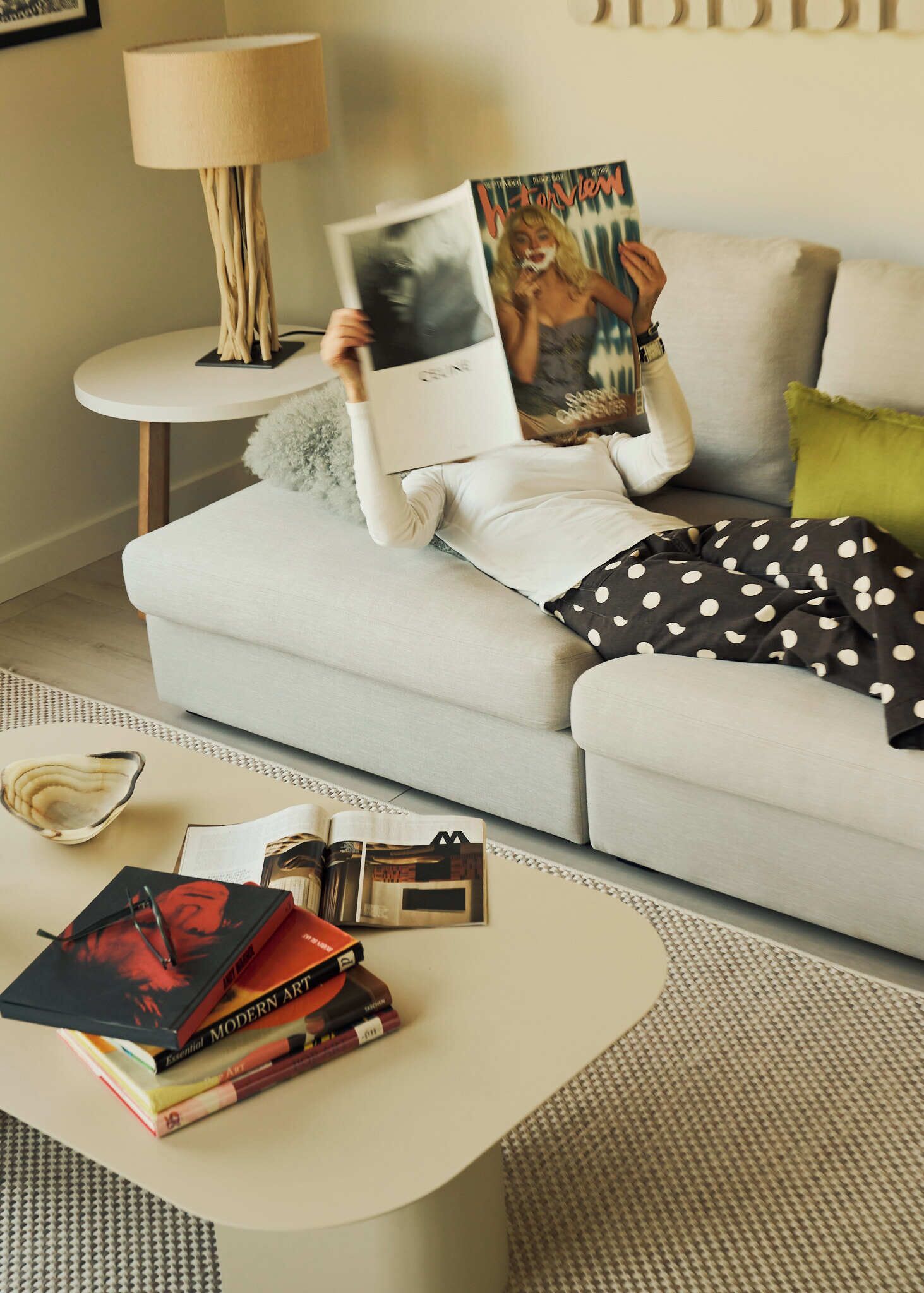 A person lying on a sofa with a magazine, surrounded by books and a lamp on a side table.