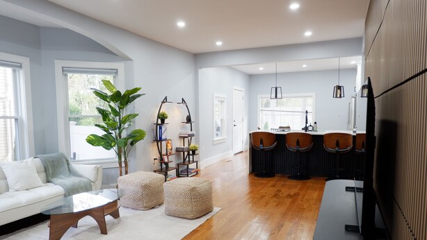 Een moderne woonkamer met een witte bank, een glazen salontafel en poufs, een eethoek met houten stoelen, en een keuken op de achtergrond.