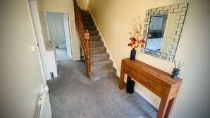 Una escalera alfombrada con barandillas de madera que conduce a un rellano. En el rellano, una consola de madera con un florero y un espejo.