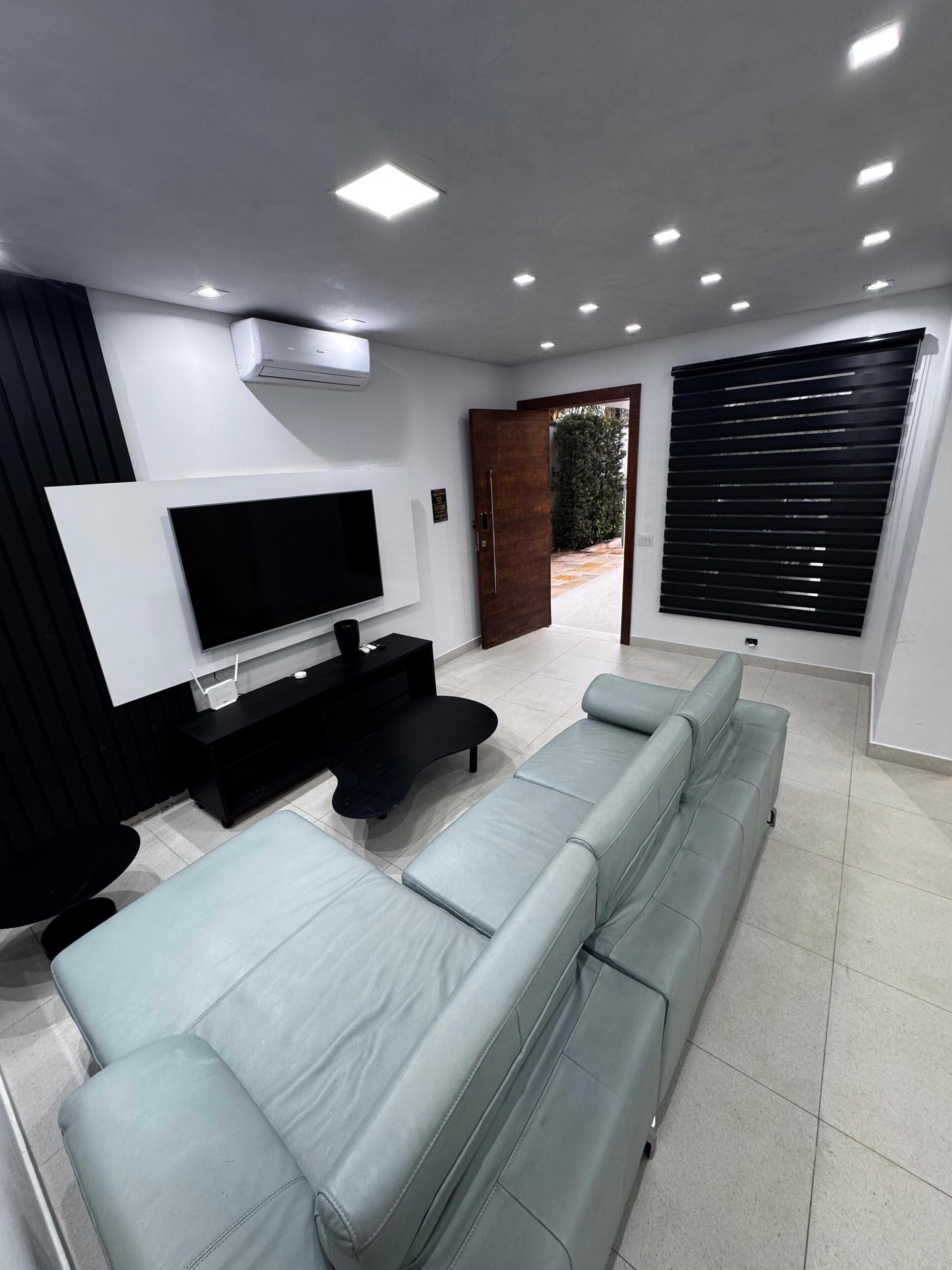Ein modernes Wohnzimmer mit Flachbildfernseher, schwarzem Unterhaltungsschrank, einem hellblauen Sofa und einem großen Fenster mit Jalousien.