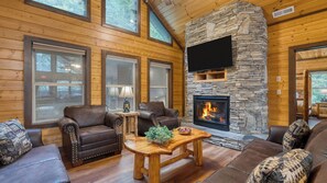 A cozy cabin interior with a stone fireplace, leather furniture, and large windows. - 16+ Guests Indoor Pool, Theater & Game Room in the Smokies Buckskin by Avantstay (Sevierville)