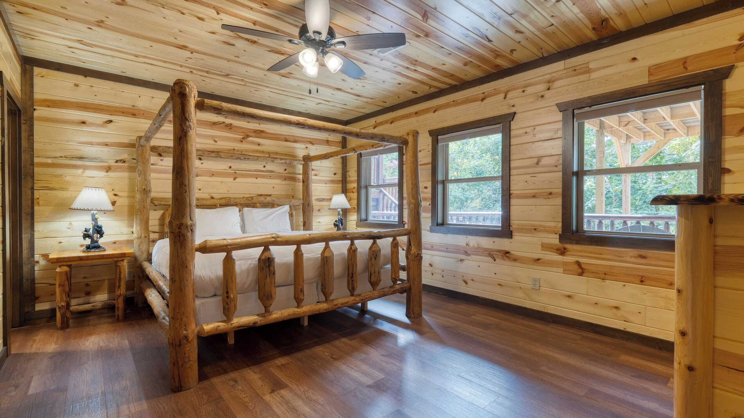 A log cabin bedroom with a four-poster bed, two bedside lamps, and large windows offering views of the outdoors.