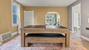 A room with a ping pong table, beige walls, a patterned rug, and a view into another room with a dining area.