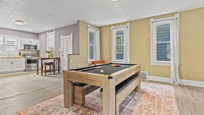 A pool table in a room with a kitchen area, a dining table with chairs, and a patterned rug.
