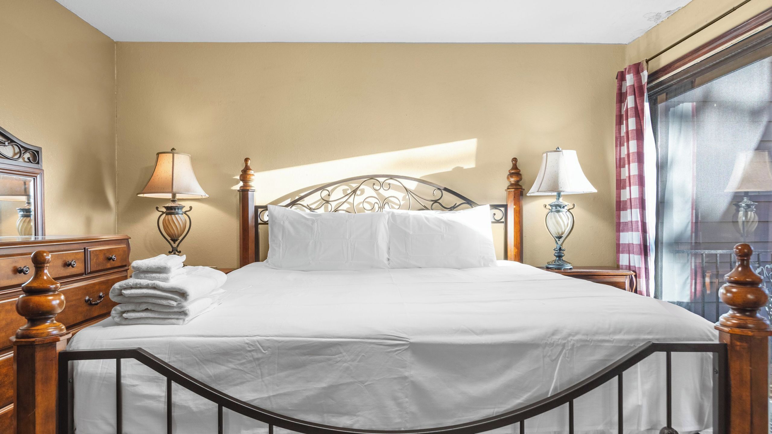 Una cama bien tendida con sábanas blancas, dos lámparas de noche, un cabecero de madera y una cómoda con espejo.
