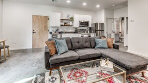 Ein modernes Wohnzimmer mit einem grauen Ecksofa, einem Glastisch und einem Teppich mit Muster. Im Hintergrund ist eine Küche mit weißen Schränken und Edelstahlgeräten zu sehen.