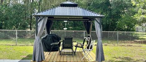 A gazebo with a metal roof and curtains, a grill, and folding chairs on a wooden deck.