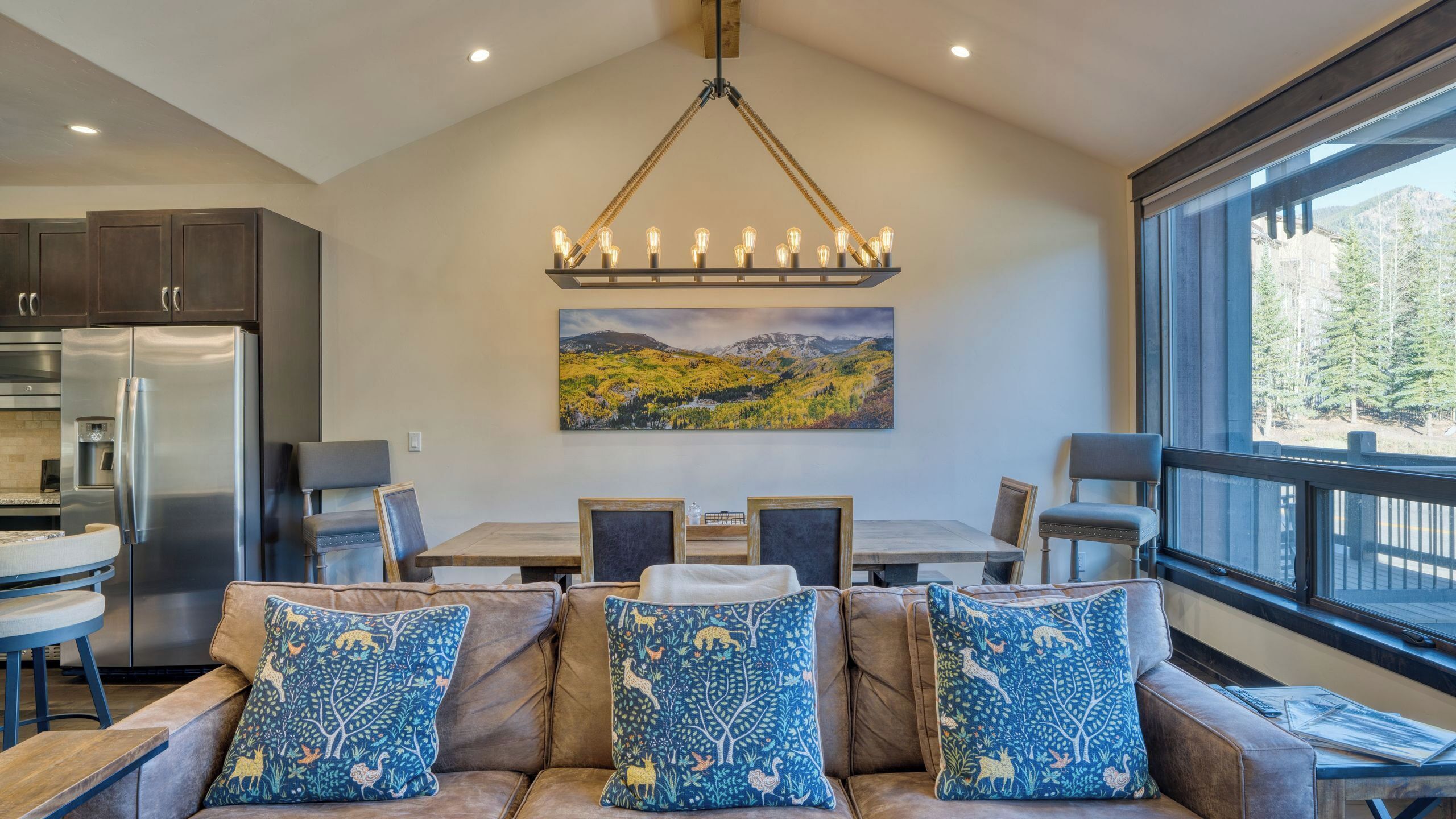 Une cuisine moderne équipée d’appareils électroménagers en acier inoxydable, un coin repas avec une table et des chaises en bois, et une grande fenêtre offrant une vue sur les montagnes.