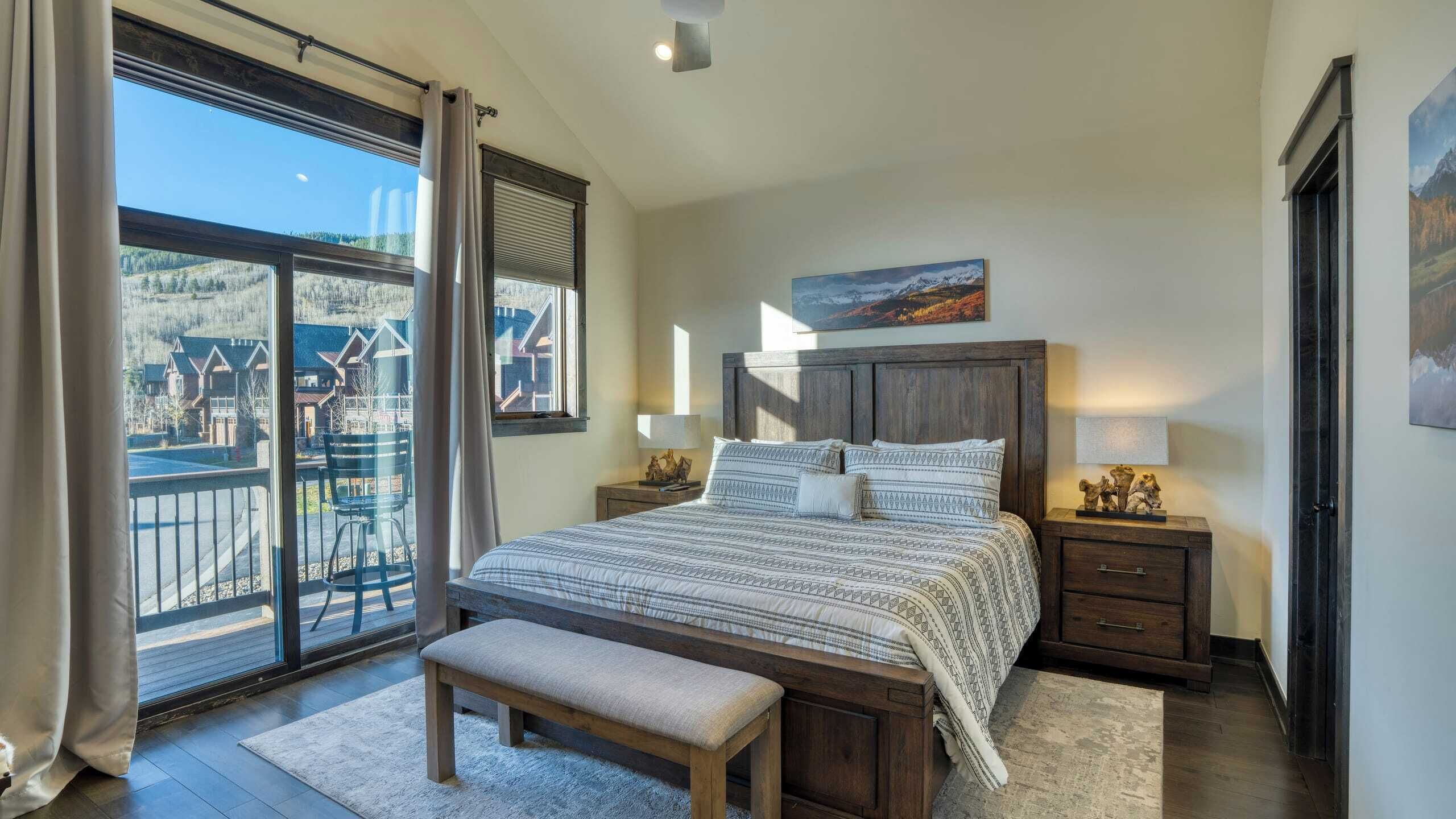 A bedroom with a large bed, a bench, a nightstand, and a view of a residential area through a sliding door.