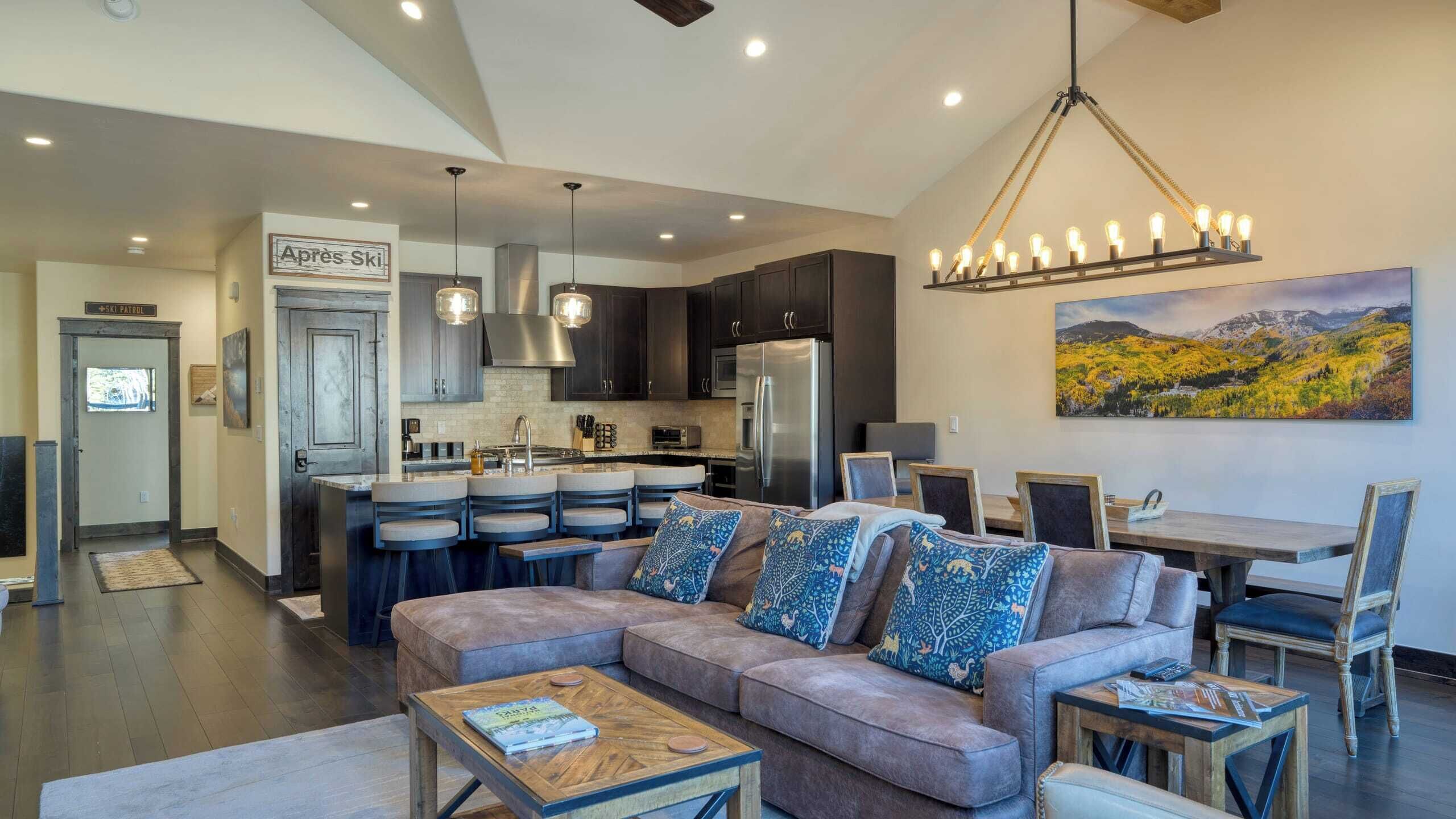 A modern living room with a sofa, coffee table, and a dining area with a table and chairs. There is a kitchen with dark cabinets and a refrigerator. A ski patrol sign is visible in the hallway.