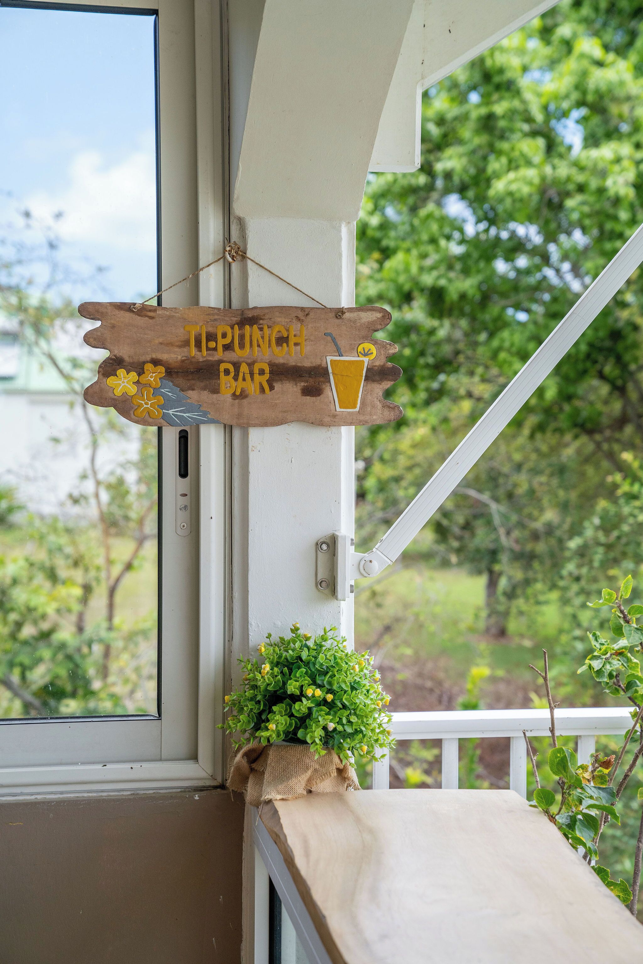 Un letrero de madera que dice "TI-PUNCH BAR" colgado cerca de una ventana con vista a la vegetación.