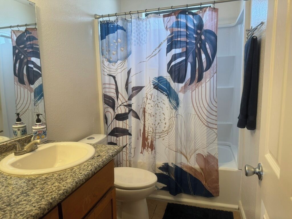 Une salle de bain équipée d’un lavabo, de toilettes et d’un rideau de douche orné d’un motif de feuilles tropicales.