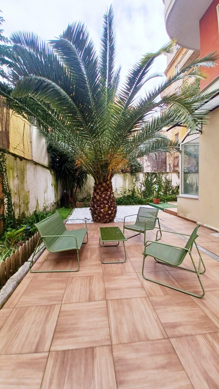 A patio with a palm tree, green chairs, and a tiled floor.
