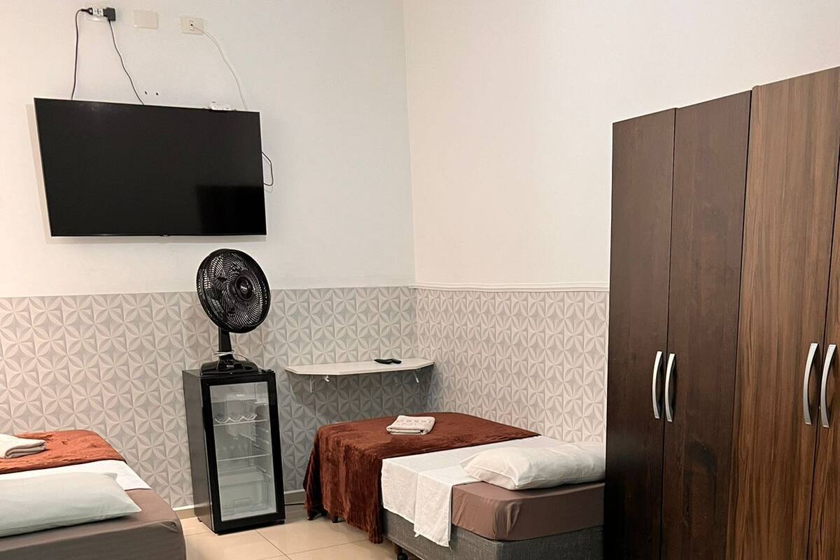 A hotel room with two beds, a wardrobe, a TV mounted on the wall, and a small table with a fan.