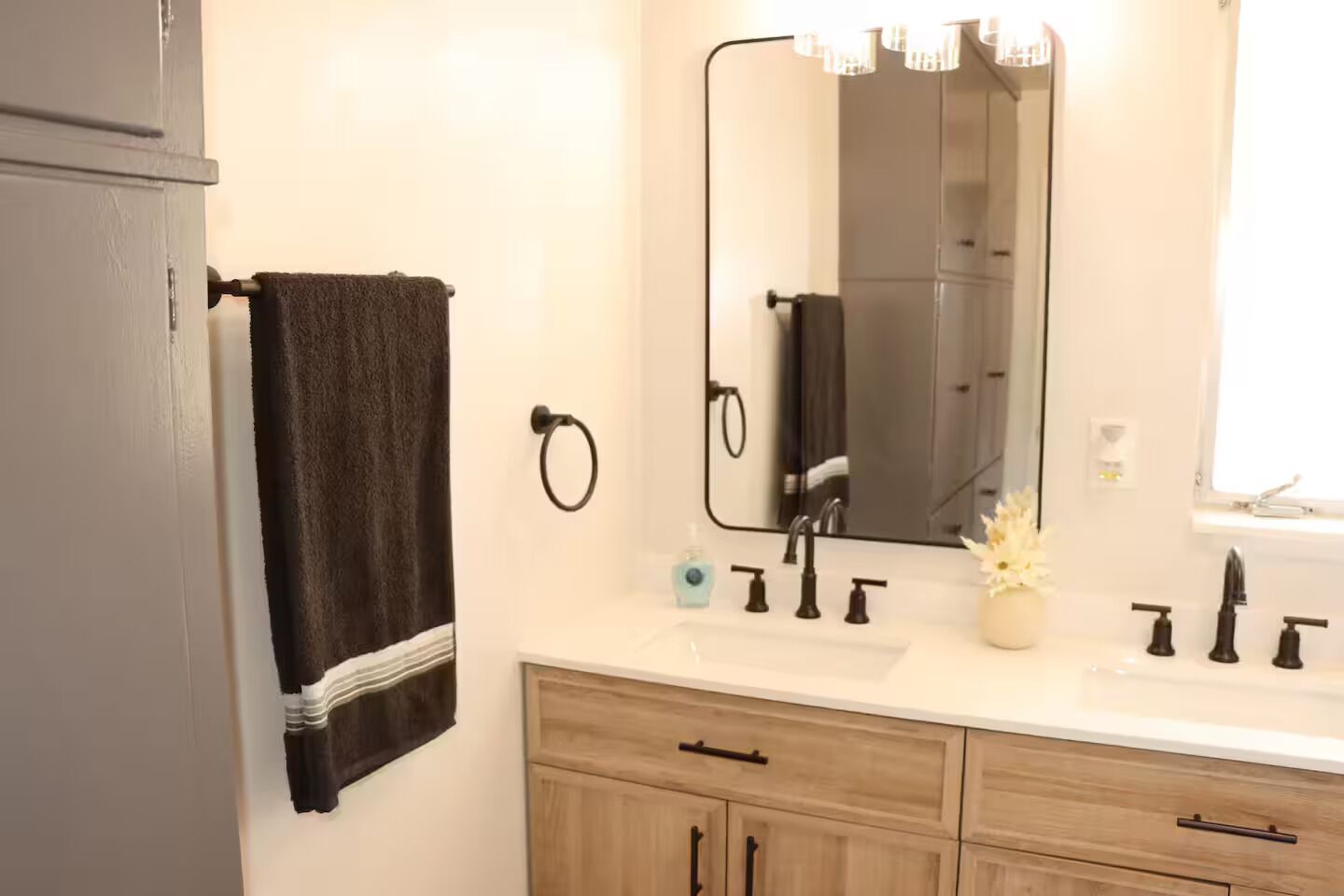 Baño con lavamanos blanco, grifo negro y un espejo encima. Hay una toalla colgada en un perchero y un mueble de madera debajo del lavamanos.