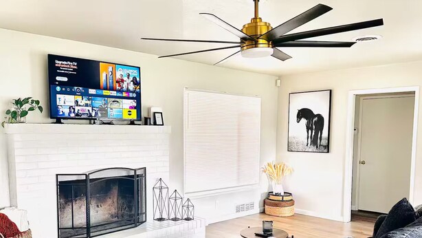 Een moderne woonkamer met een open haard, een grote grijze bank, een plafondventilator en een tv aan de muur die een sportzender toont.
