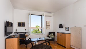 A modern living room with a glass-top dining table, black chairs, a black rug, a wooden cabinet, a television, and a window with a view of greenery. - One Bedroom Apartment with Loggia and Sea View (Dubrovnik)