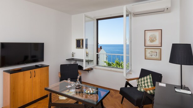 A modern living room with a large flat-screen TV, a glass-top dining table, and a view of the sea through an open window. - One Bedroom Apartment with Loggia and Sea View (Dubrovnik)