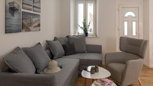 Ein modernes Wohnzimmer mit einem grauen Ecksofa, einem kleinen runden Tisch und einem grauen Sessel. An der Wand hängen drei Gemälde, ein Fenster mit Blick ins Freie und eine Tür mit rundem Sturz.