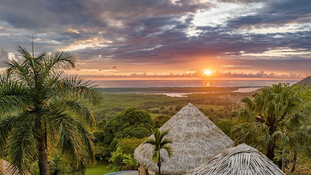 Une construction à toit de chaume avec une piscine, entourée d’une végétation luxuriante et offrant une vue pittoresque sur le coucher du soleil.