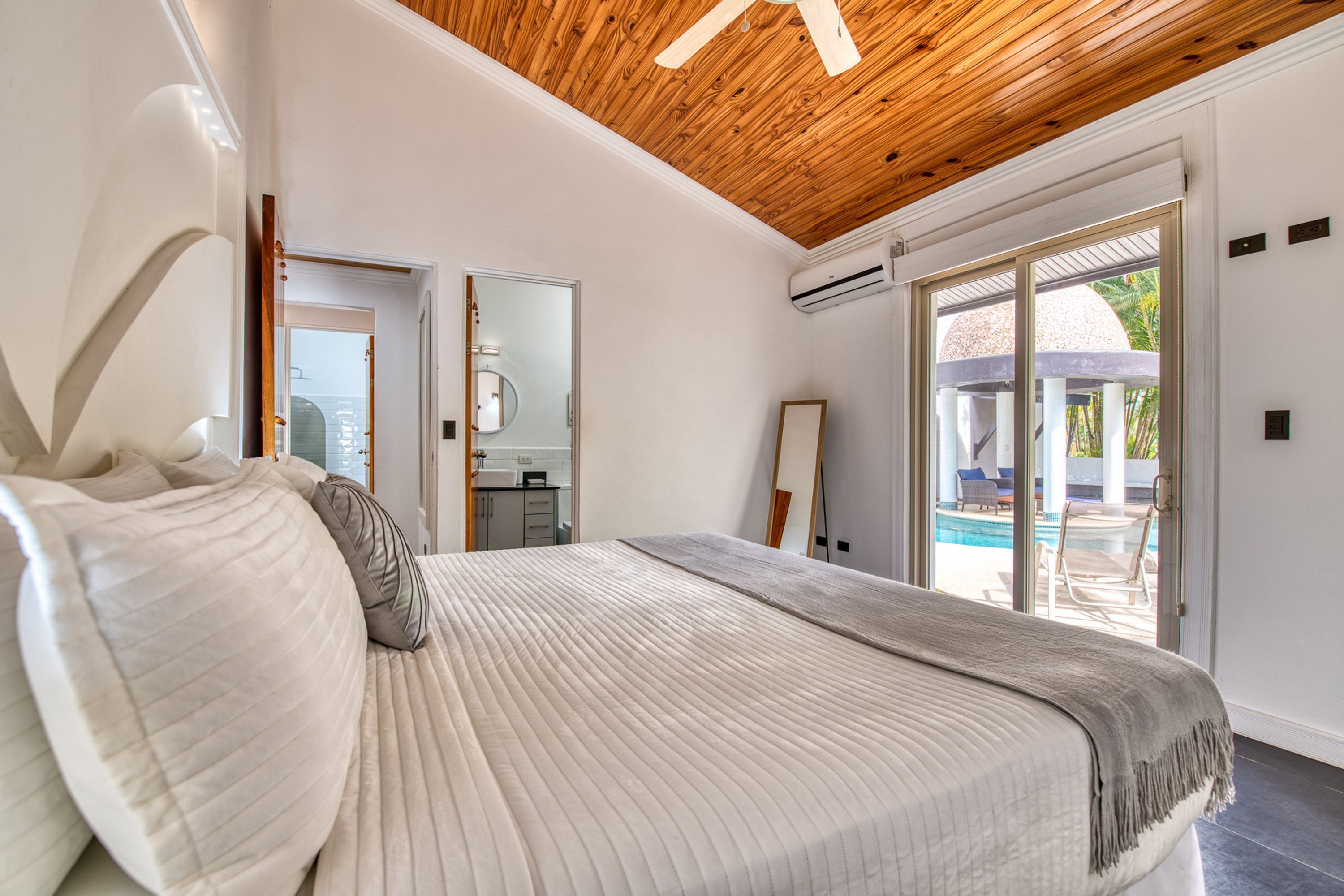 A bedroom with a large bed, wooden ceiling, and a view of an outdoor pool area.