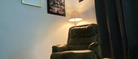 Una habitación con un sillón reclinable, un cartel enmarcado de un jugador de baloncesto y una lámpara sobre una mesa.