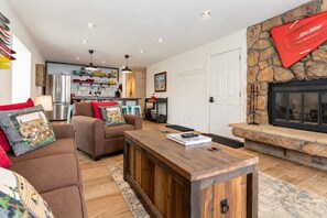 A modern living room with a fireplace, a kitchen in the background, and a stone accent wall. - Charming 2BR Fraser Escape – Fireplace, Trails & Resort Nearby! (Fraser)