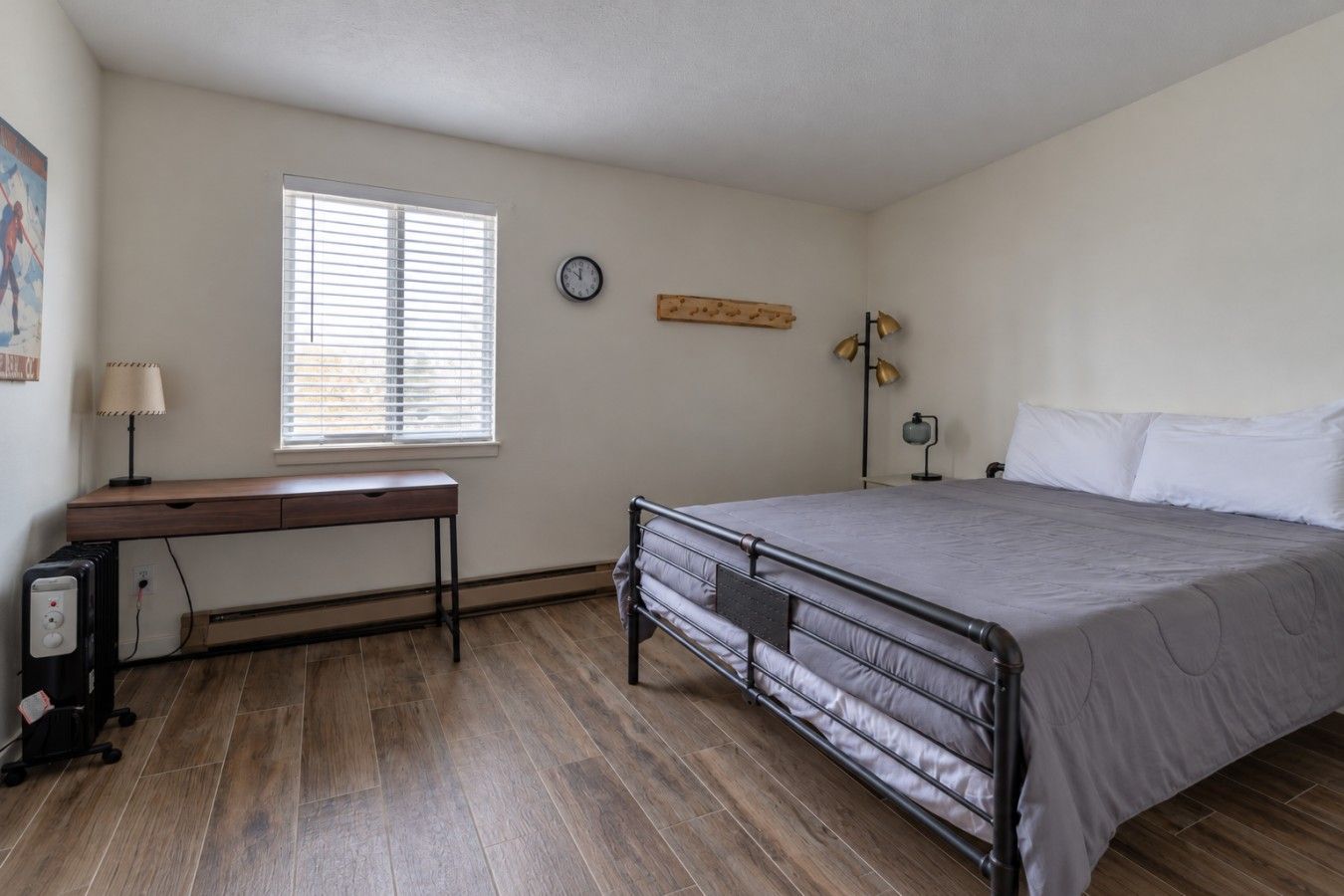 A bedroom with a bed, a desk, a chair, a clock, and a window with blinds.