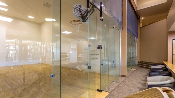 Una cancha de baloncesto moderna en interior con paredes de vidrio, suelo de madera y una zona de asientos con bancos acolchados.