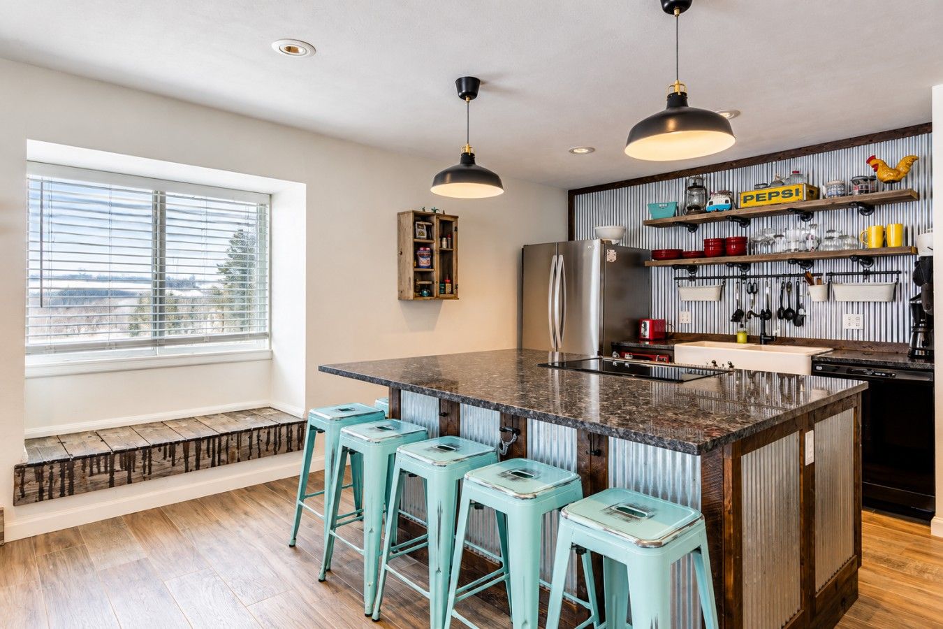 Una cocina con una isla central, taburetes de bar, un refrigerador y un ventanal con persianas.