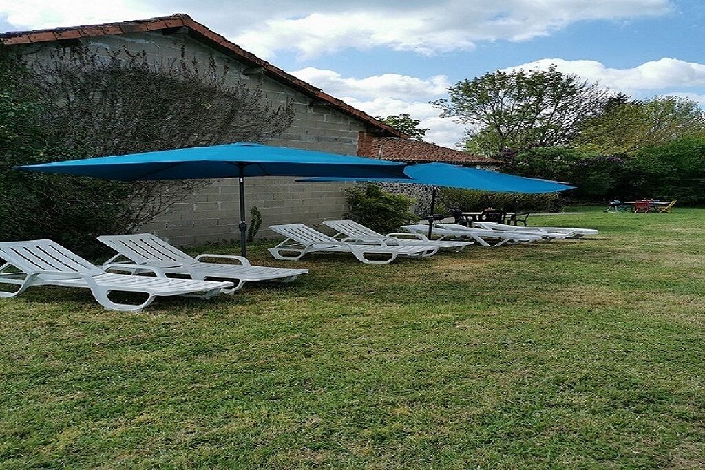 A row of white lounge chairs under a blue umbrella, a brick building, and a grassy area.