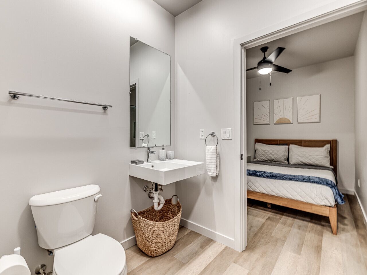 Une salle de bain moderne avec des toilettes, un lavabo et un miroir. La chambre adjacente est équipée d’un lit en bois avec une tête de lit et d’un ventilateur de plafond.