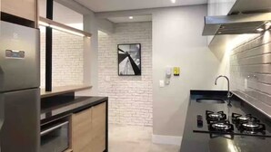 A modern kitchen with a stainless steel refrigerator, wooden cabinets, a black countertop, a gas stove, and a brick wall backsplash. - Itaim Star 2 (São Paulo)