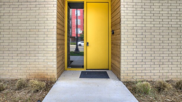 A yellow door with a glass panel, set in a brick wall with a concrete step and a doormat. - Midtown Luxe 1 Bed Apt (Oklahoma City)