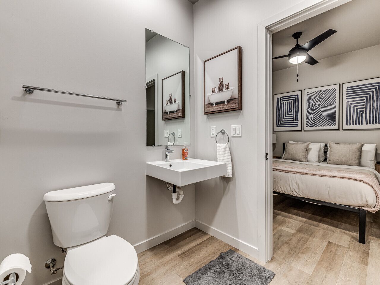 Un baño moderno con inodoro, lavabo y espejo. En la habitación contigua, hay una cama con cabecera y un ventilador de techo.