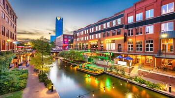 A riverside walkway with buildings on either side, a boat docked on the river, and a tall blue building in the background.