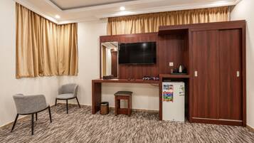 A hotel room with a desk, two chairs, a television, a small refrigerator, and a trash bin.