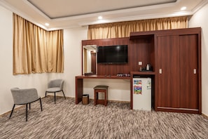 Una habitación de hotel con un escritorio, dos sillas, un televisor, un refrigerador pequeño y un cubo de basura.