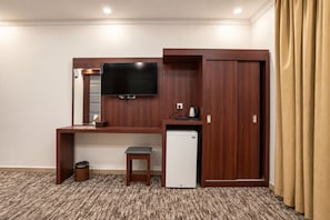 A hotel room with a wooden desk, a television, a mini-fridge, a trash bin, and a wardrobe. - MG Crown Hotel 1 (Madinah)