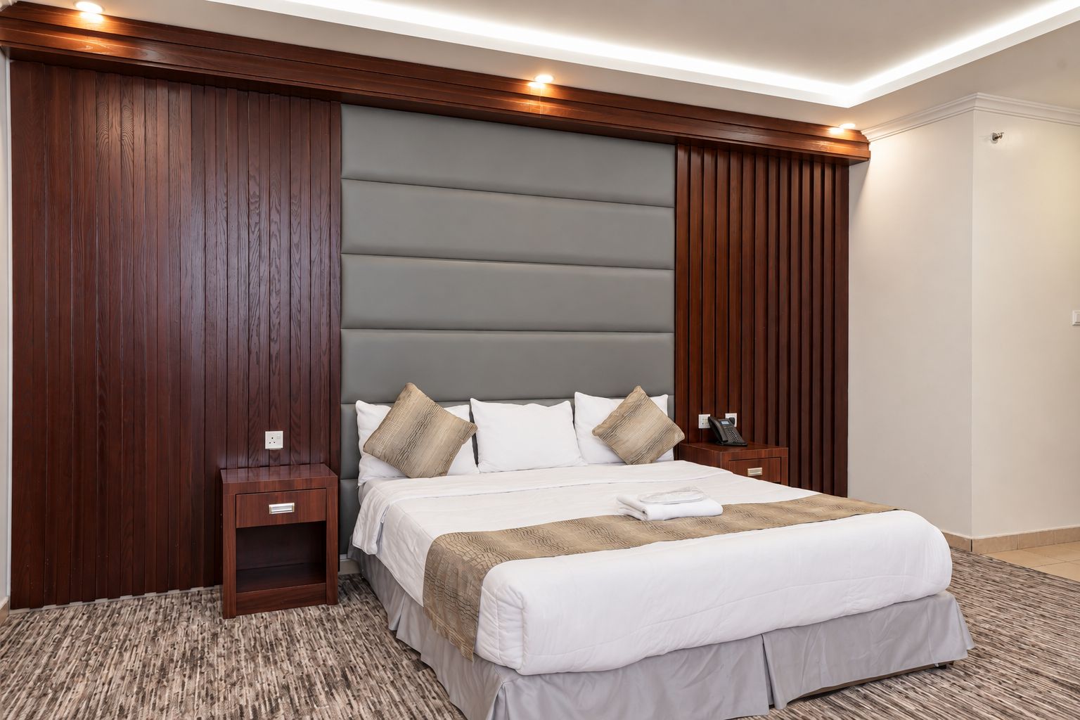 Habitación de hotel con una cama grande, paneles de madera, un cabecero acolchado gris y una mesita de noche pequeña con un teléfono.