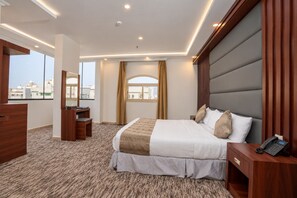 A hotel room with a large bed, a desk, a chair, a mirror, and a view of the city. - MG Crown Hotel 1 (Madinah)