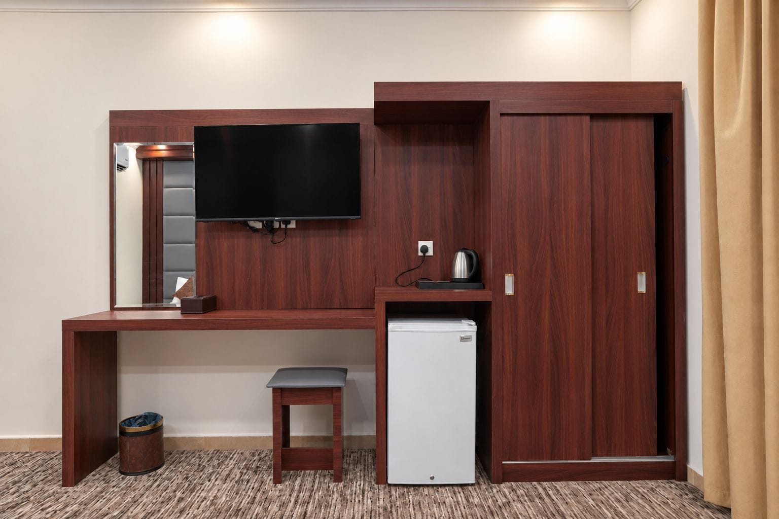 Een compacte hotelkamer met een houten bureau, een flatscreen televisie, een minibar, een klein krukje en een prullenbak.