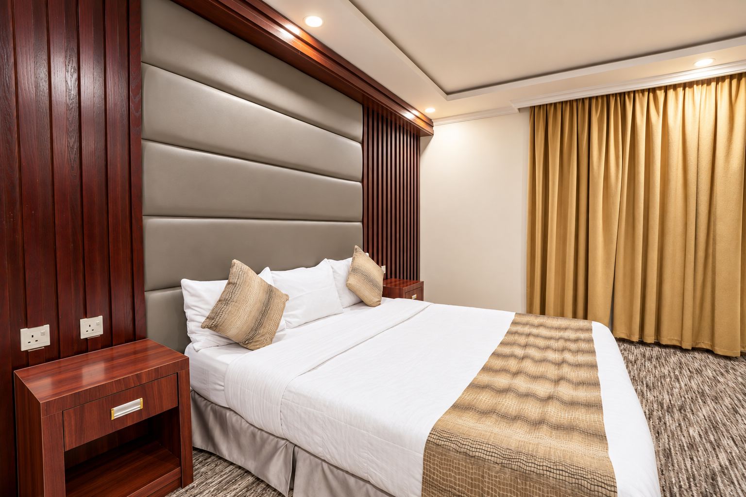 Ein Hotelzimmer mit einem großen Bett, einem Nachttisch aus Holz, einem an der Wand befestigten Kopfteil und einem Fenster mit Vorhängen.