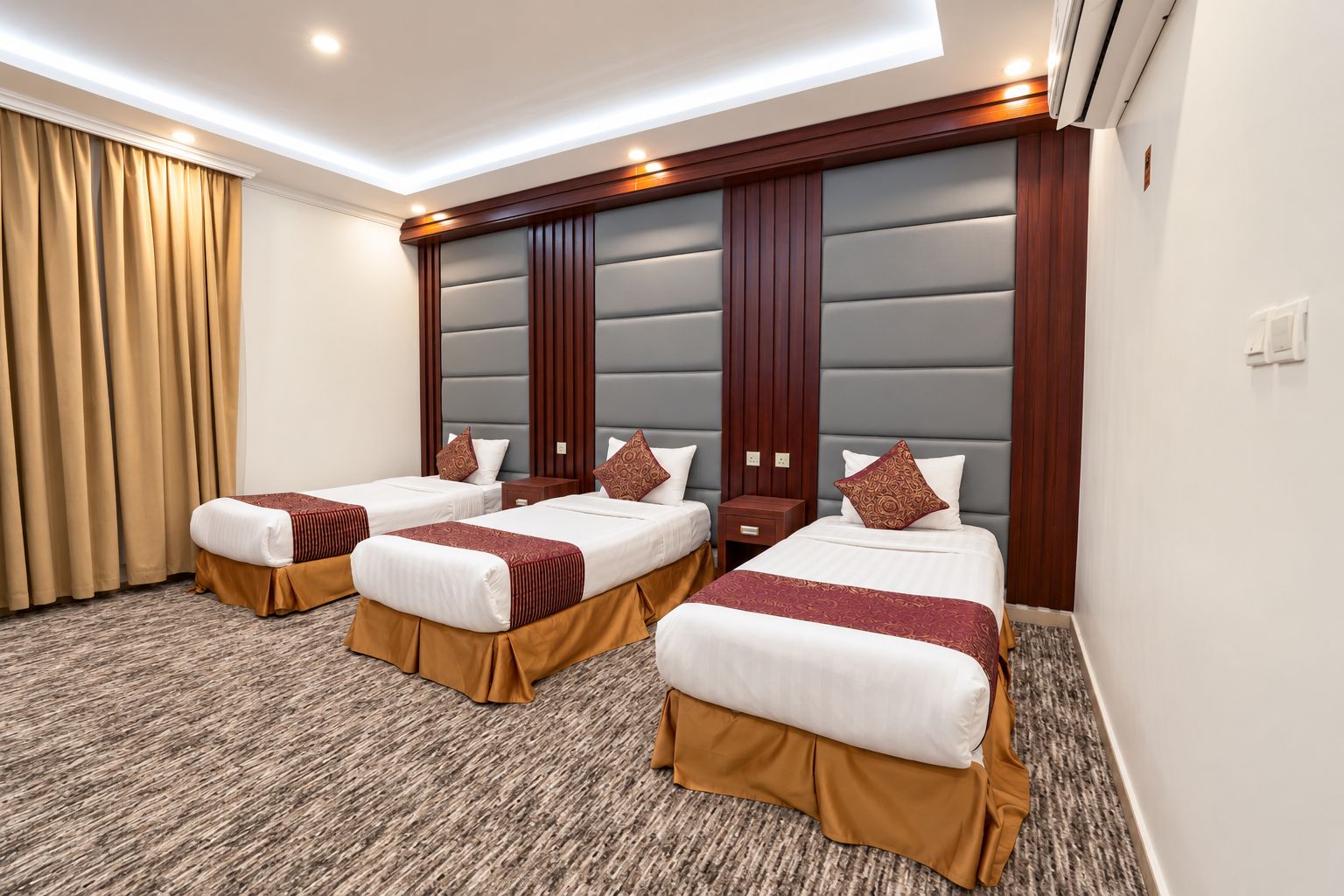 Een hotelkamer met drie bedden, elk met een bruine stoffen onderrok, een witte matras en een bedsprei met dessin. De kamer heeft een groot gestoffeerd hoofdeinde, een vloerbedekking en een raam met gordijnen.